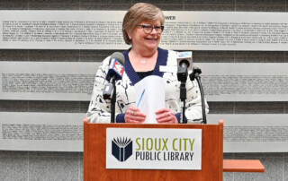 A woman stands at a podium labeled Sioux City Public Library, smiling and holding papers. Microphones are attached to the podium, and a large wall with names is in the background.