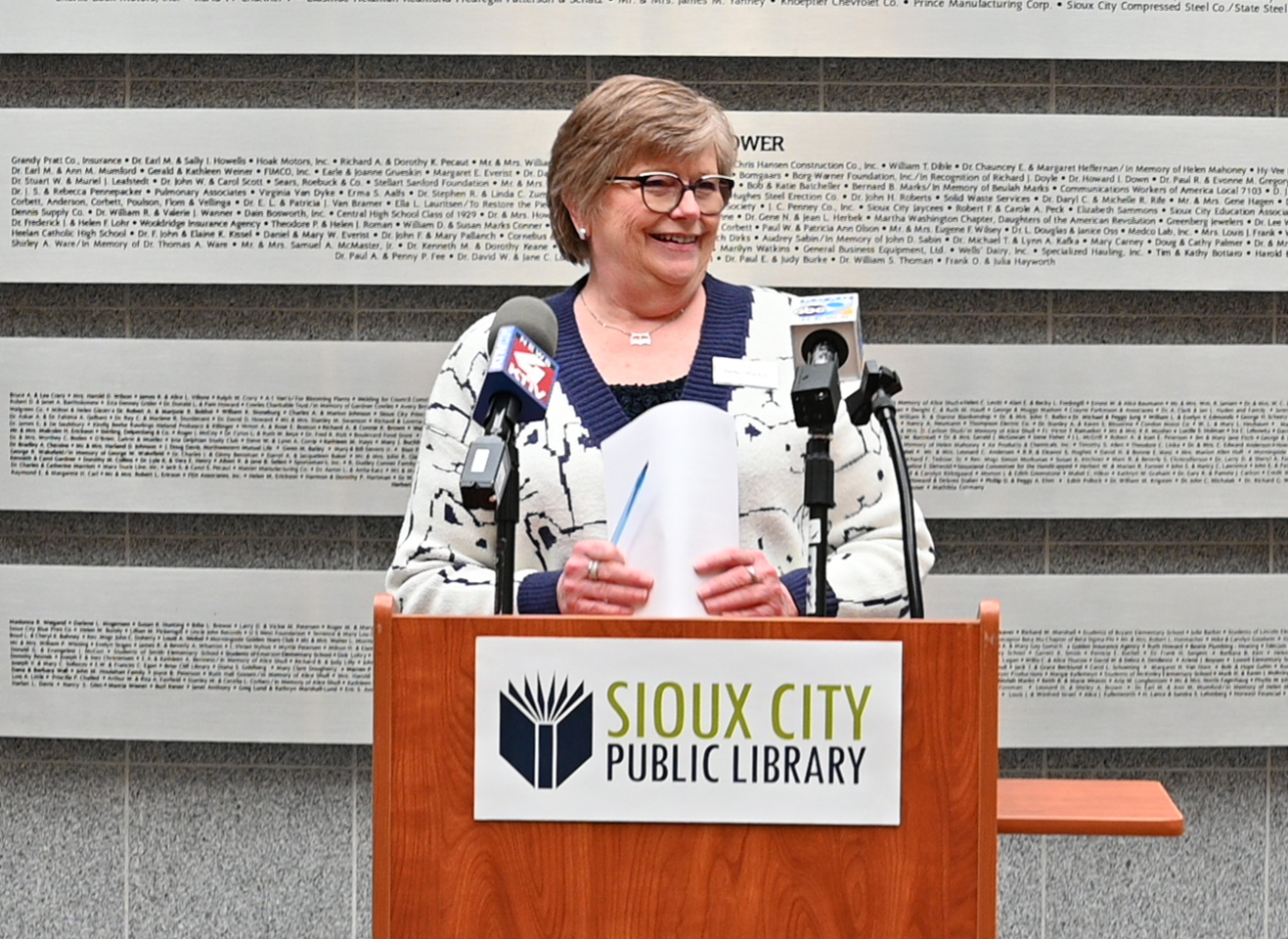 A woman stands at a podium labeled Sioux City Public Library, smiling and holding papers. Microphones are attached to the podium, and a large wall with names is in the background.