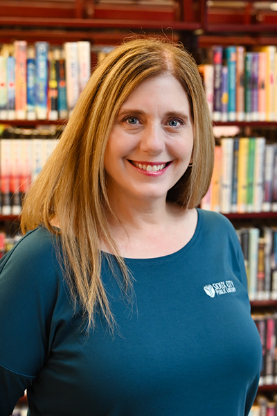 A woman with long light brown hair, wearing a teal shirt with a Sioux City Public Library logo, smiles while standing in front of bookshelves filled with colorful books.